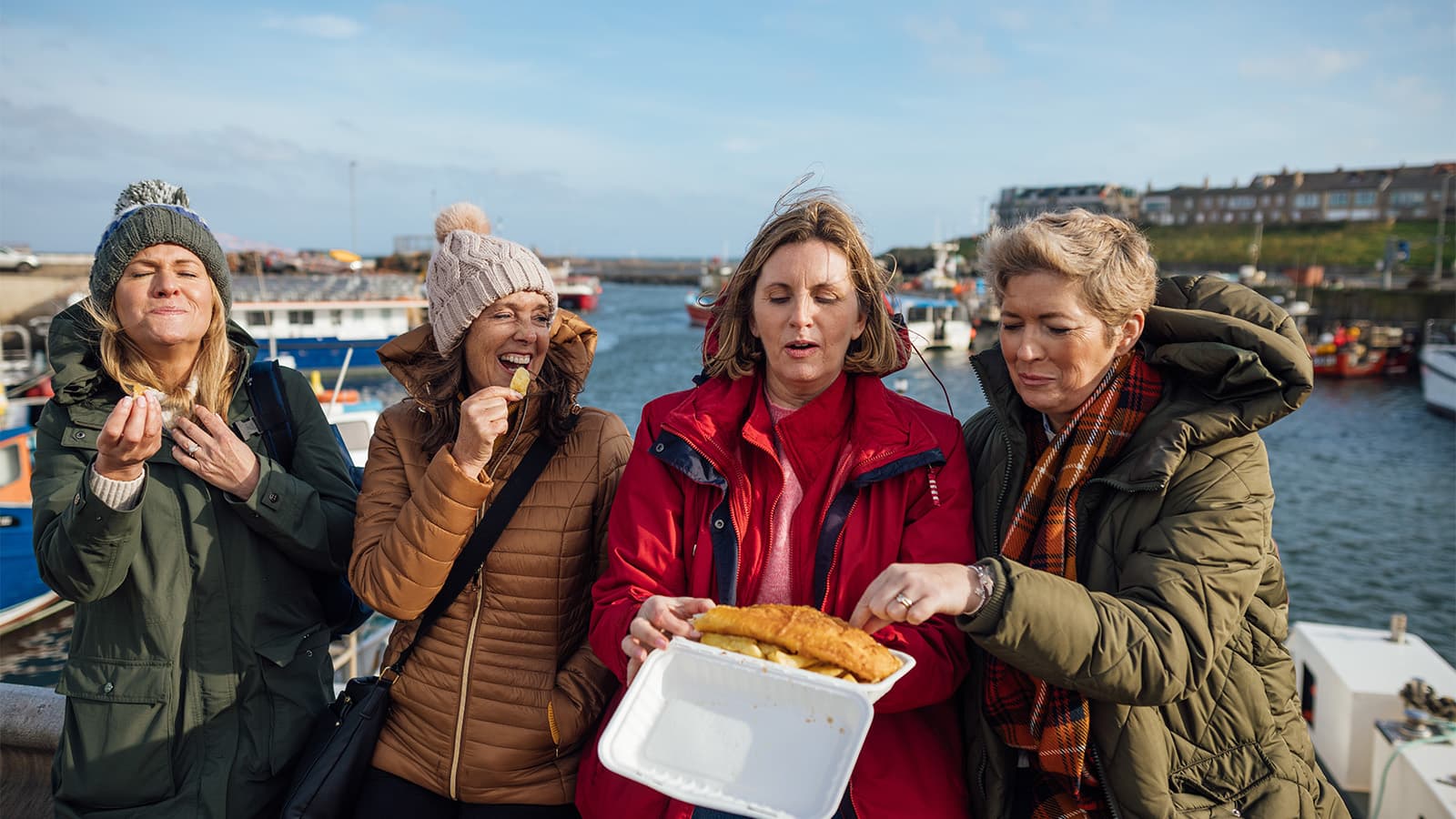 Women at the seaside sharing fish and chips while earing autumnal clothing in the cold