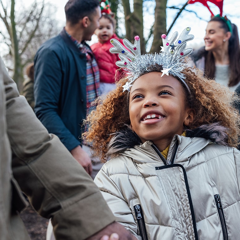 hero image for winter gifting: family outdoors, girl in a shiny silver warm winter coat wearing both tinsel on her head and a celebratory (brithday?) tiara. There is a family in the background bokeh blur