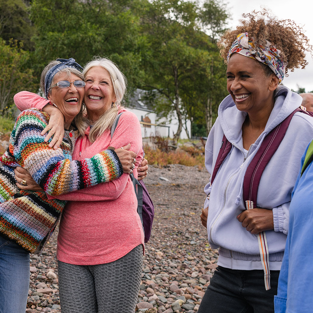 A group of people hiking on a rocky beach with trees and a house in the background. Two women are embracing warmly on the left, while others stand nearby wearing casual clothing and backpacks.